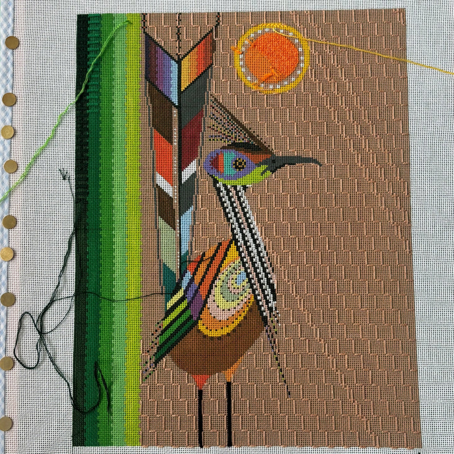 image showing a needlepoint canvas pinned to a frame. On the canvas there is a bird painted with many colored feathers and a sun in the background