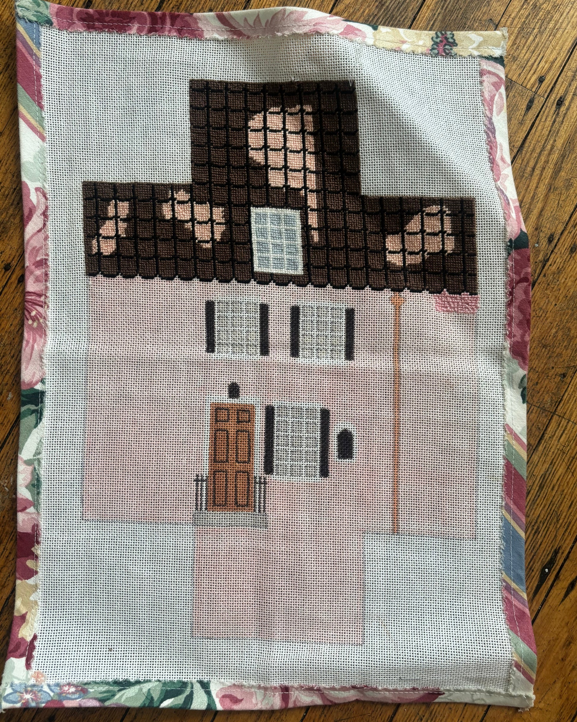 An image of a needlepoint brick cover in the style of a pink house with a brown roof. 