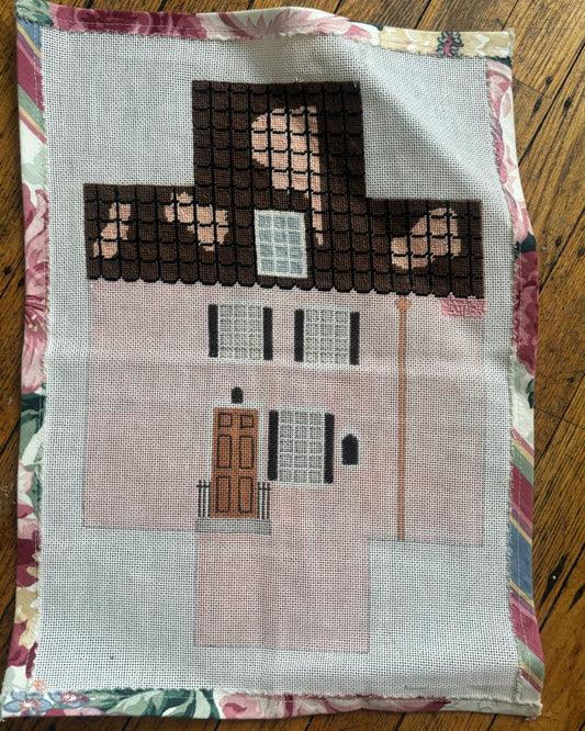 An image of a needlepoint brick cover in the style of a pink house with a brown roof. 